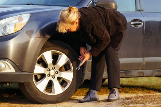 Unrecognizable blonde woman is leaning over her car and changing broken tire on sunny autumn day in nature. Tourism and travel. Adventures and vacations. Strong independent women.