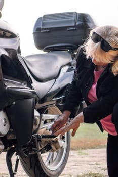 Unrecognizable mature blonde repairs wheel of her motorcycle at stop on way in nature in autumn with wrench. Hobby motorcycle tourism. Maintenance of motorcycles. Female mechanic. Tourism and travel.