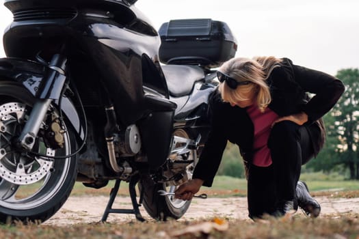 Middle-aged woman is kneeling by wheel of her bike and repairing it on autumn day on road.Hobby motorcycle tourism.Maintenance . Female mechanic. Excellent physical shape. Tourism travel.