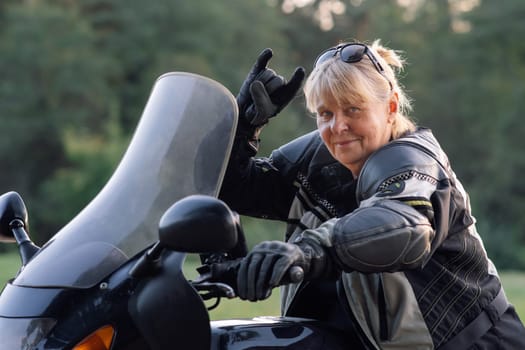 Super mom, blonde biker granny gestures ok while sitting on her motorcycle. Hobby motorcycle tourism. Healthy lifestyle. Emotional relief. Excellent physical shape. Tourism and travel.