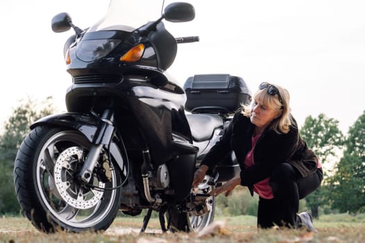 Mature blonde woman in leather jacket repairs wheel of her motorcycle at stop on way to nature in autumn. Hobby motorcycle tourism. Healthy lifestyle. Excellent physical shape. Tourism travel.