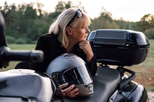 Motorcycle tourism. Mature blonde stands near her motorcycle on autumn day and looks away. Healthy lifestyle. Excellent physical shape. Motorcycle equipment. Hobbies, travel, communication.