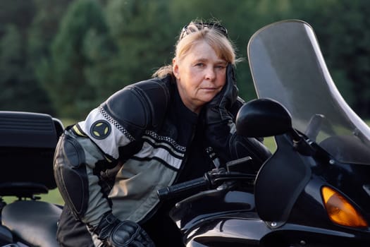 Mature woman in leather gear on a black motorcycle in nature in autumn during parking. Motorcycle tourism. Healthy . Sport strength of character. Excellent physical shape. Motorcycle equipment.