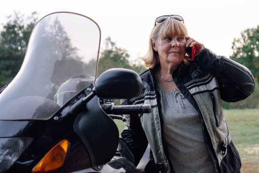 Modern stylish confident women. Attractive happy mature blonde biker is talking on red phone near black motorcycle. Hobby - motorcycle tourism. Female biker. Tourism and travel.