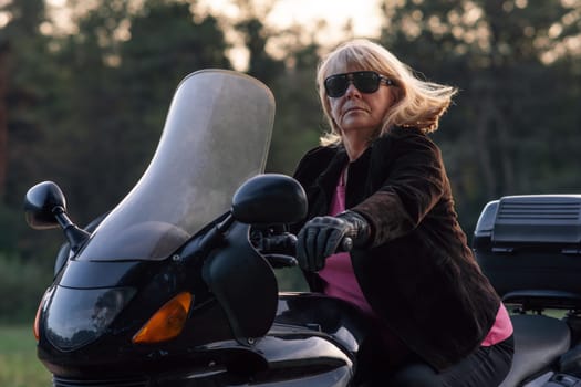 Stylish middle-aged blonde motorcyclist in leather jacket, black gloves is getting ready for road on autumn day. Hobby. Healthy lifestyle. Emotional relief. Excellent physical shape.Tourism travel.
