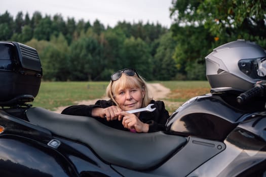Serene mature blonde with wrench looks into camera near her bike against background of rural road stretching into distance on autumn day. Hobby - motorcycle tourism. Maintenance .Tourism and travel.
