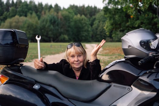No problem , female mechanic with wrench gestures near black motorcycle on rural road on autumn day. Modern strong confident women. Hobby - motorcycle tourism. Mature female mechanic and biker.