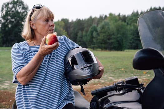 Female biker. Eating apple pretty blonde middle-aged biker near her bike at bus stop during motorcycle rally. Hobbies of strong independent women. Tourism d travel. Adventures. Motorcycle tourism.