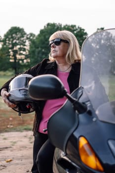 Vertical portrait of mature blonde in jacket with helmet in her hands standing near motorcycle in autumn in nature. Motorcycle tourism. Healthy lifestyle. Sport and strength of character.