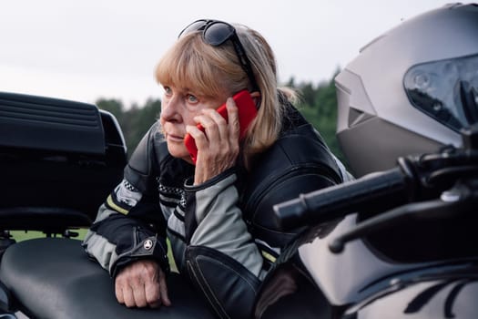 Tired middle-aged biker woman is talking on red phone about her black motorcycle on autumn day on road. Hobby - motorcycle tourism. Female biker. Communication in travel. Mobile communications.