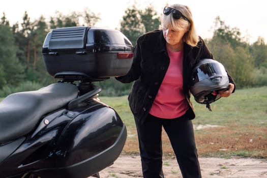 Pretty mature blonde in black trousers, black jacket with motorcycle helmet under her arm puts something in trunk of motorcycle. Hobby motorcycle tourism. Healthy lifestyle.Tourism and travel.