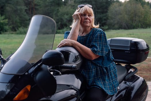 Hobby - motorcycle tourism. Pretty mature blonde in blue blouse looks thoughtfully to side, sitting on her motorcycle. Female biker. Tourism and travel. Strong independent women. Adventures .