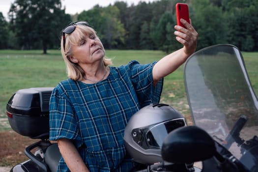 Attractive mature woman in dark blue plaid blouse sits on motorcycle and takes selfie with her phone. Hobby - motorcycle tourism. Female biker. Tourism travel. Strong independent women. Adventures.