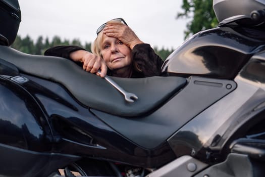 Thoughtfulness and fatigue are shown by mature female mechanic withwrench near black motorcycle on rural road on autumn day. Modern confident women. Hobby - motorcycle tourism. Tourism and travel.