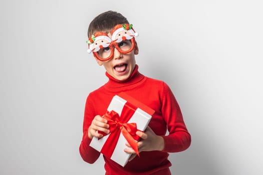 Smiling little boy wearing funny glasses in form of Christmas Santa Claus holding Xmas gift while standing in a white background, dressed in warm red xmas sweater.