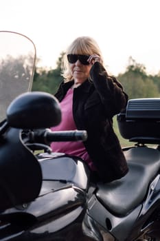 Portrait of mature blonde motorcyclist in black glasses in leather jacket, sitting on her bike on autumn day. Hobby . Healthy lifestyle. Emotional relief. Excellent physical shape. Tourism travel.