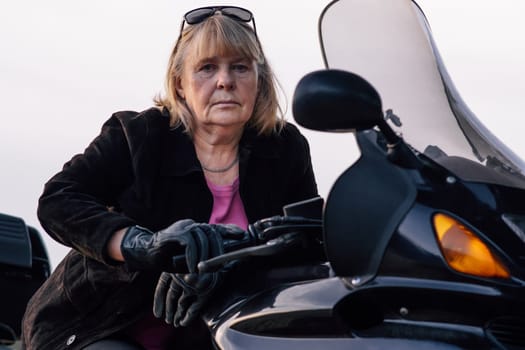 Seriously looking at camera adult blonde female motorcycle tourist in black gloves, sitting on her bike. Motorcycle tourism. Healthy lifestyle. Excellent physical shape. Hobbies, travel .