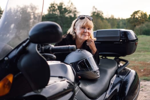 Positive emotions. Smiling happily, middle-aged blonde looks into camera, leaning on her black bike. Hobby motorcycle tourism. Healthy lifestyle. Excellent physical shape. Tourism and travel.