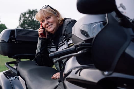Mobile communications. Charming smiling middle-aged biker woman talking on phone sitting on her motorcycle on autumn day on road. Hobby - motorcycle tourism. Female biker. Communication in travel.