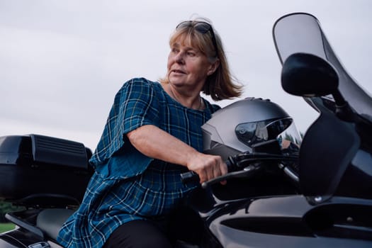Female biker. Independent mature blonde is having great time, looking away while sitting on her black motorcycle on autumn day in nature. Hobby - motorcycle tourism. Tourism and travel. Adventures.