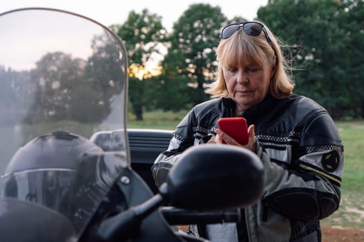 Pretty blonde middle-aged biker is dialing number on her red iPhone, standing next to motorcycle in nature. Hobby - motorcycle tourism. Female biker. Communication in travel. Mobile communications.