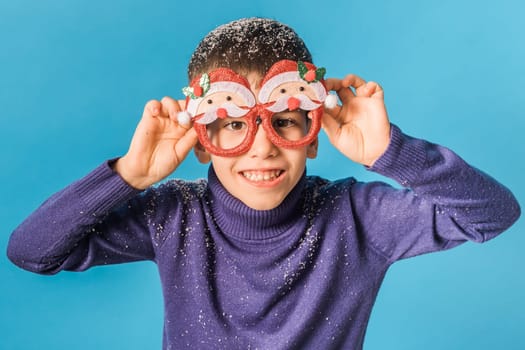 Portrait of attractive cheerful boy funny wearing festive santa eyeglasses eve christmas time isolated over bright blue color background.