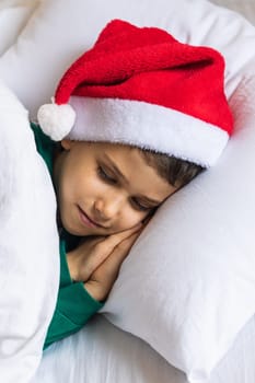 Sweet child boy sleeping in bed wearing Santa Claus hat in Christmas eve.