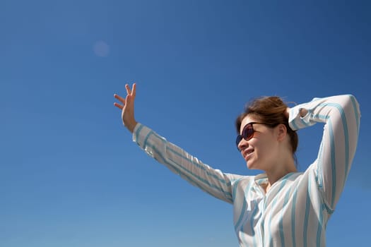 happy blonde in glasses and a light shirt waving her hand to someone on a background of blue sky. High quality photo