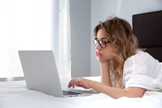 A beautiful girl with glasses is lying on a white blanket in the morning light, chatting, online, typing on a computer keyboard - the concept of a social network, online shopping.