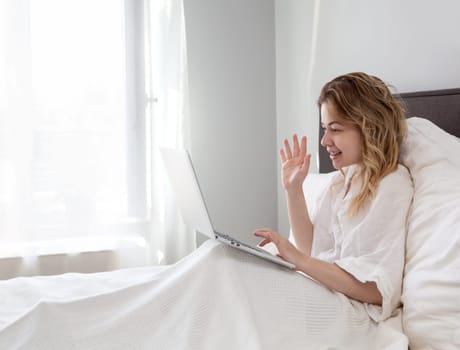 Beautiful girl sitting in a white bed in the morning light, chatting, online networking, freelancer, business, online shopping, social network concept. High quality photo