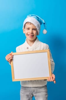 Child boy in blue santa hat and blank canvas. Copy space on canvas board for image or message. Kid in Christmas hat holds mockup poster and standing over blue background. Generation alpha and gen alpha.