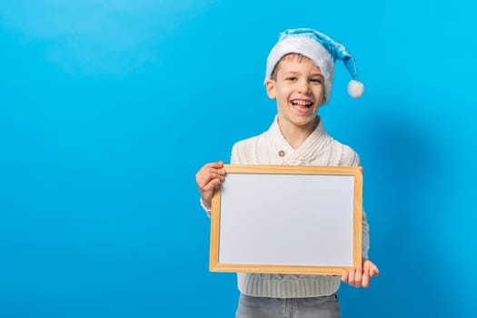 Child boy in blue santa hat and blank canvas. Copy space on canvas board for image or message. Kid in Christmas hat holds mockup poster and standing over blue background. Generation alpha and gen alpha.