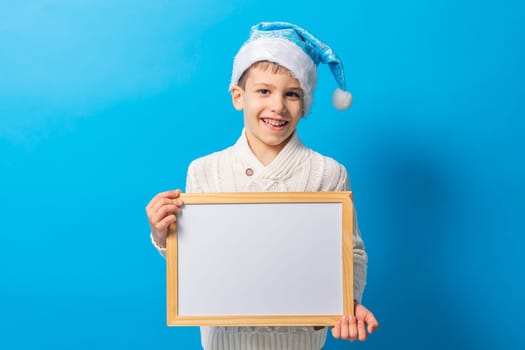 Child boy in blue santa hat and blank canvas. Copy space on canvas board for image or message. Kid in Christmas hat holds mockup poster and standing over blue background. Generation alpha and gen alpha.