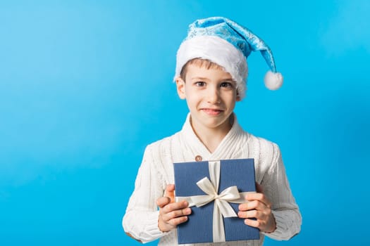 Happy little boy in blue Christmas hat with a gift box copy space. Empty place for your text. Winter holidays and present concept