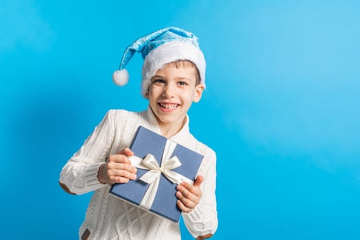 Happy little boy in blue Christmas hat with a gift box copy space. Empty place for your text. Winter holidays and present concept