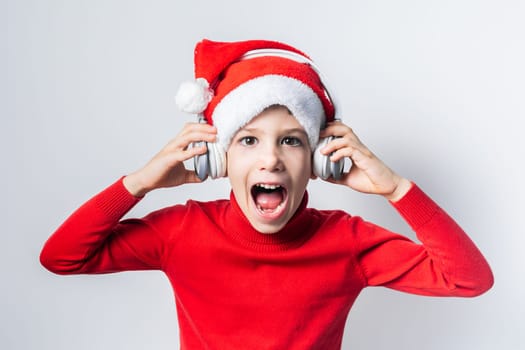 Funny child boy holds listening to Christmas music with Santa Claus hat on white background. Generation alpha and gen alpha children