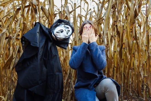 Woman covering her face with her hands near creepy scarecrow with mask of death in cornfield on Halloween.