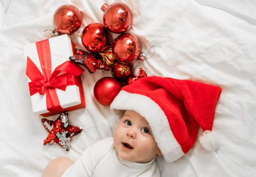 Christmas toddler in Santa hat lying on bed with gift box and festive baubles top view.