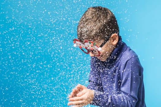 Child boy blowing snow out of his hands on blue background. Christmas and New Year holidays concept. Generation alpha and gen alpha children