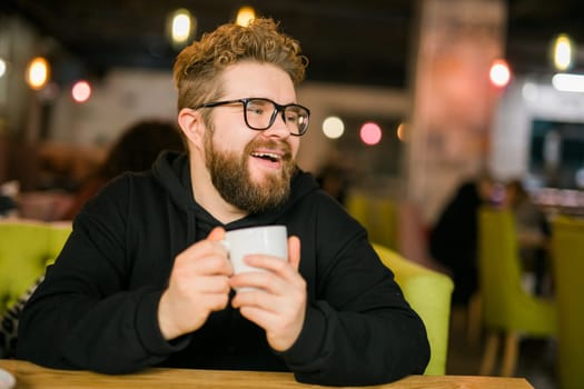 Bearded curly man smiling confident drinking coffee in restaurant or coffee shop. Millennial generation and Gen Y.