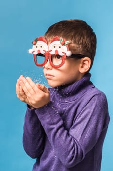 Child boy blowing snow out of his hands on blue background. Christmas and New Year holidays concept. Generation alpha and gen alpha children