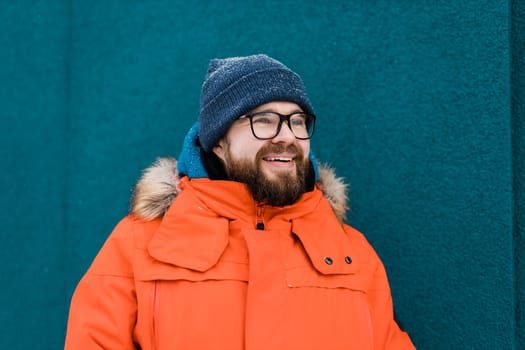 Portrait handsome eyeglasses young male smiling bearded stands on a blue wall background in a bright orange winter jacket with a hood with fur in winter. Millennial generation and Gen Y