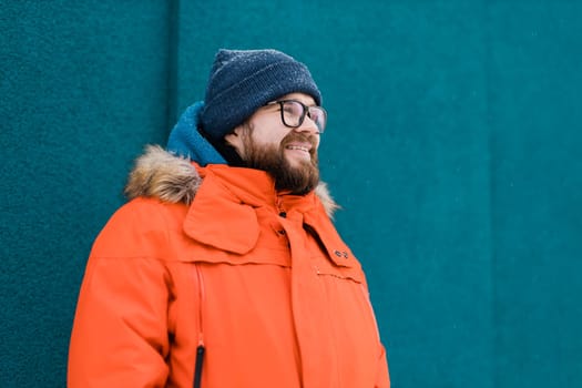 Portrait handsome eyeglasses young male smiling bearded stands on a blue wall background in a bright orange winter jacket with a hood with fur in winter. Millennial generation and Gen Y