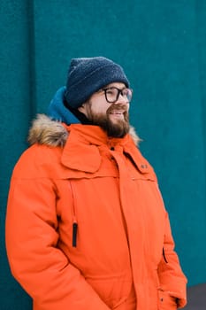 Portrait handsome eyeglasses young male smiling bearded stands on a blue wall background in a bright orange winter jacket with a hood with fur in winter. Millennial generation and Gen Y