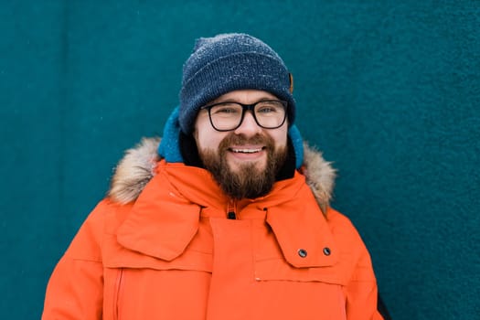 Portrait handsome eyeglasses young male smiling bearded stands on a blue wall background in a bright orange winter jacket with a hood with fur in winter. Millennial generation and Gen Y