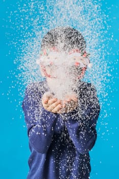 Child boy blowing snow out of his hands on blue background. Christmas and New Year holidays concept. Generation alpha and gen alpha children