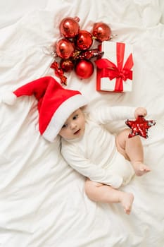 Christmas toddler in Santa hat lying on bed with gift box and festive baubles top view.