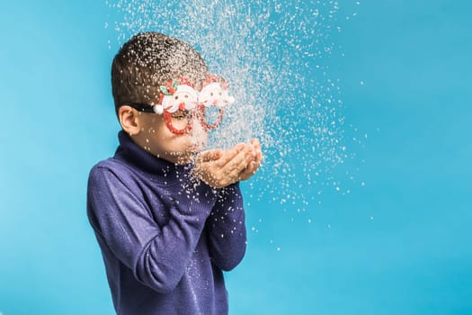 Child boy blowing snow out of his hands on blue background. Christmas and New Year holidays concept. Generation alpha and gen alpha children