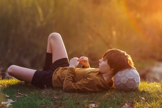 Waiting for workout in summer morning at sports camp. Teenager lies on mown grass at stadium, resting his head on soccer ball. Sports and health. Children's sports sections. Football practice.