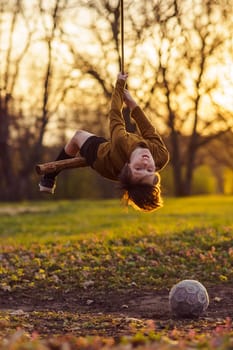 Active games outside in early autumn. Happy teenage boy is hanging upside down on homemade swing in nature, in park, in village in rays of sunset, football is lying nearby. Positive emotions.